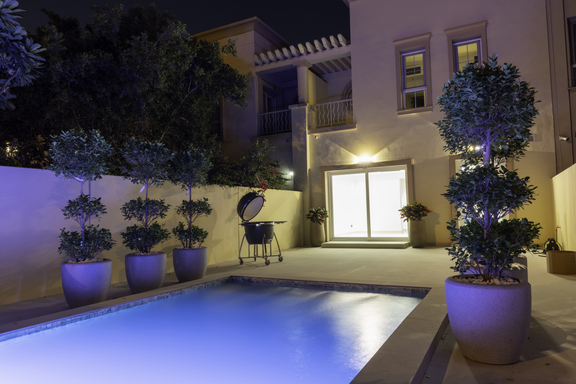 Pool terrace at night with barbecue station and planted trees against the villa facade