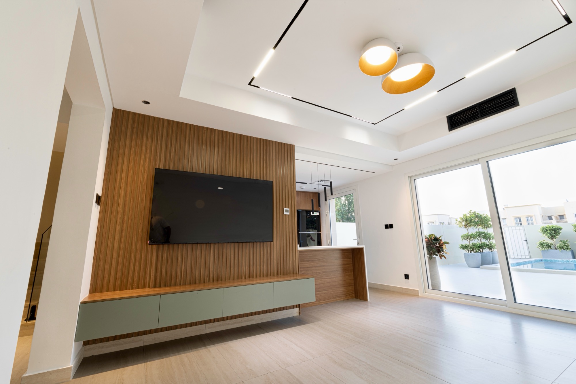 Living room with fluted walnut media wall, sage cabinetry and track lighting opening onto a pool terrace